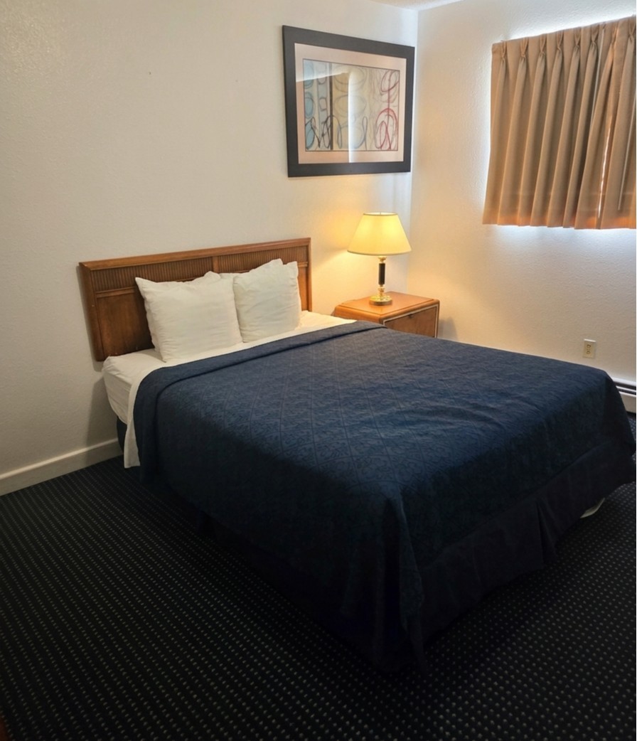 Furnished bedroom at Sunset Suites with a queen bed, navy bedspread, wood headboard, nightstand and lamp